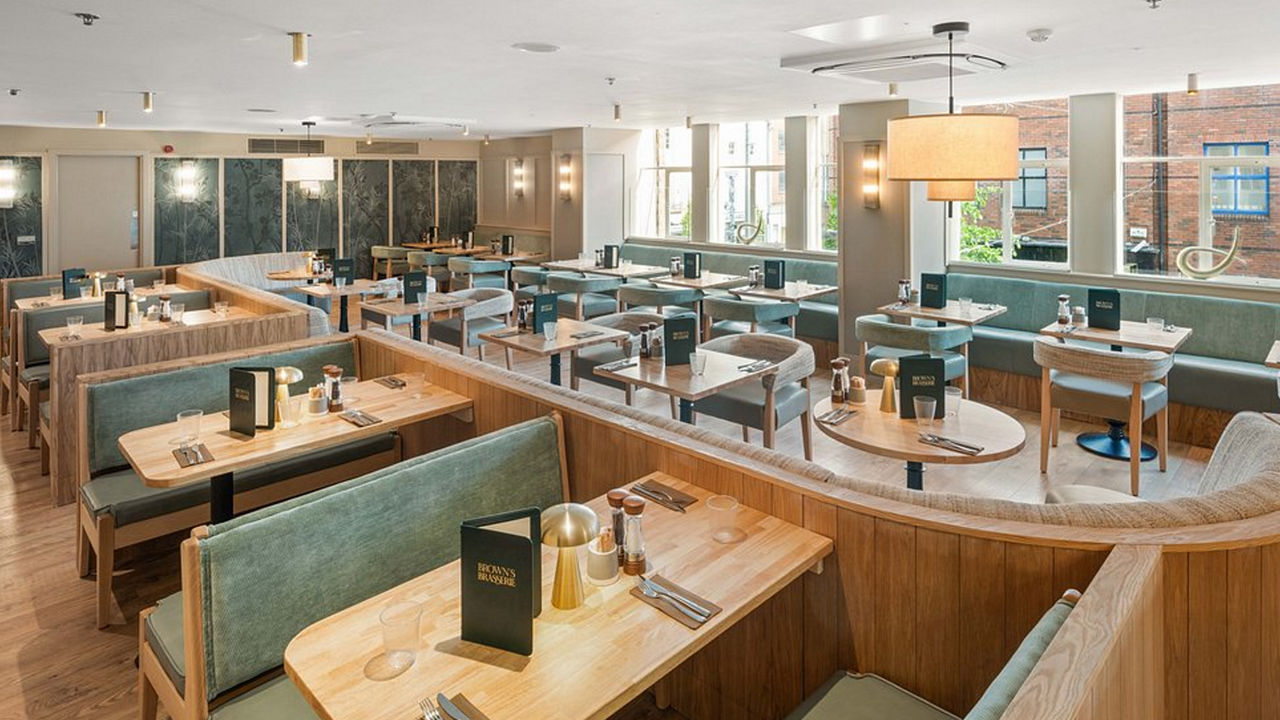 Contemporary restaurant interior with wooden booths and tables, green cushioned seating, menus on tables, and large windows.