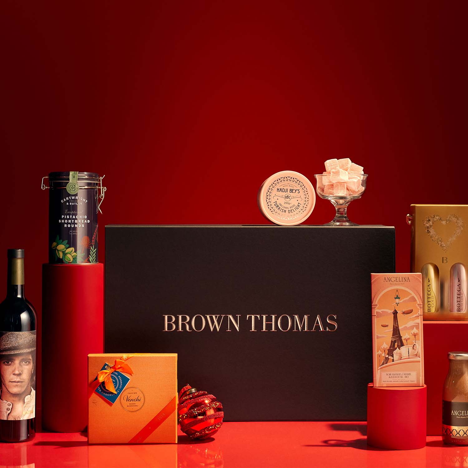 A selection of luxury gift items are arranged against a red background, featuring a prominent Brown Thomas box and various gourmet products.