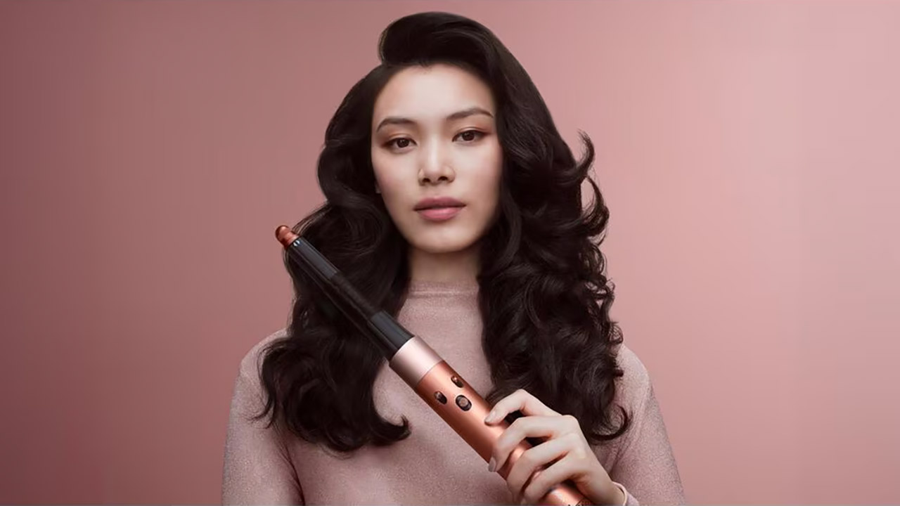 A woman with styled hair holds a Dyson curling tool against a soft pink background.
