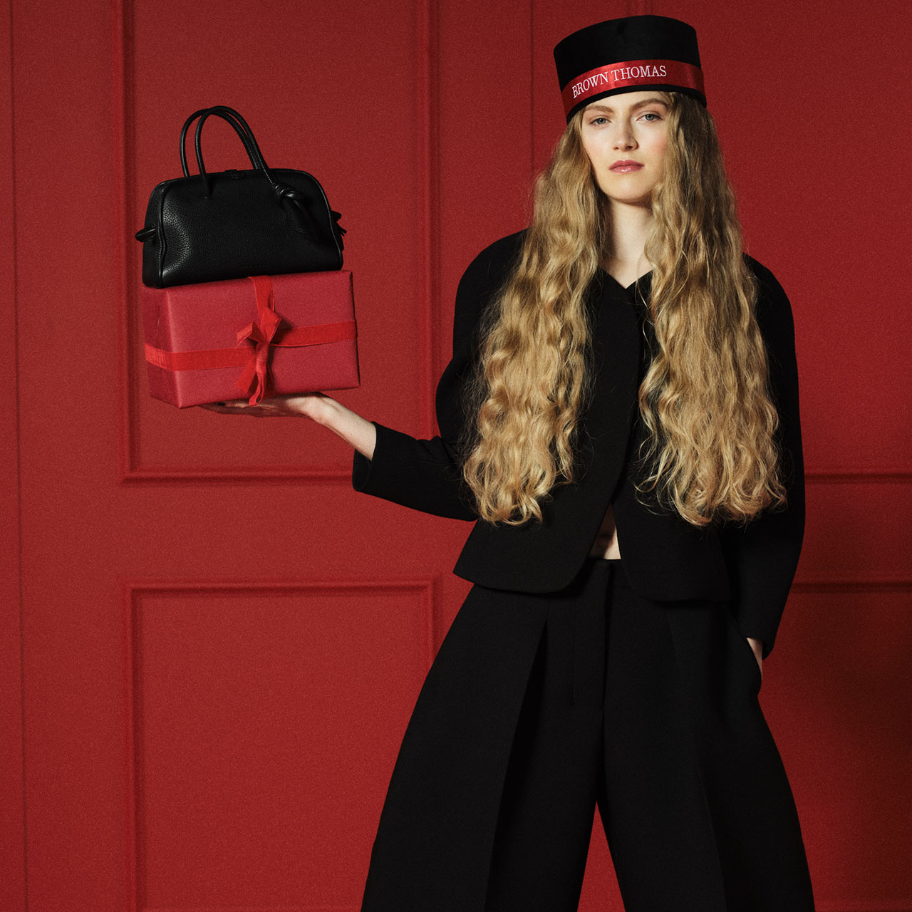 A woman with long hair, wearing a black and red hat, holds a black handbag and a red gift box against a red background.