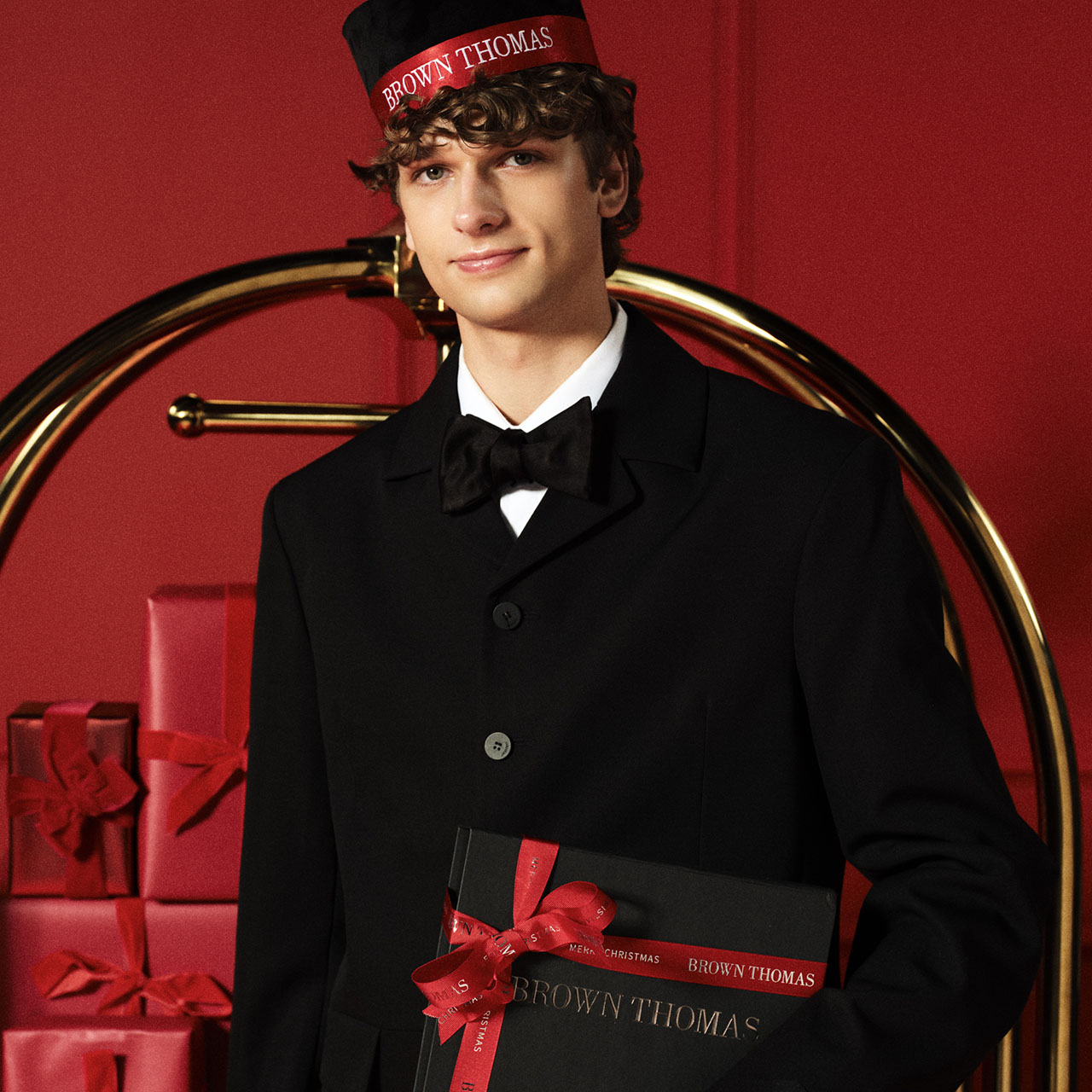A person in a formal black uniform and hat holds a gift box, standing in front of a luggage cart with more wrapped gifts.
