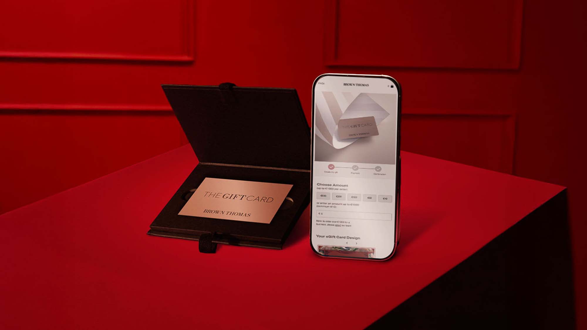 A gift card in a black folder beside a smartphone displaying gift card details, set on a red surface.