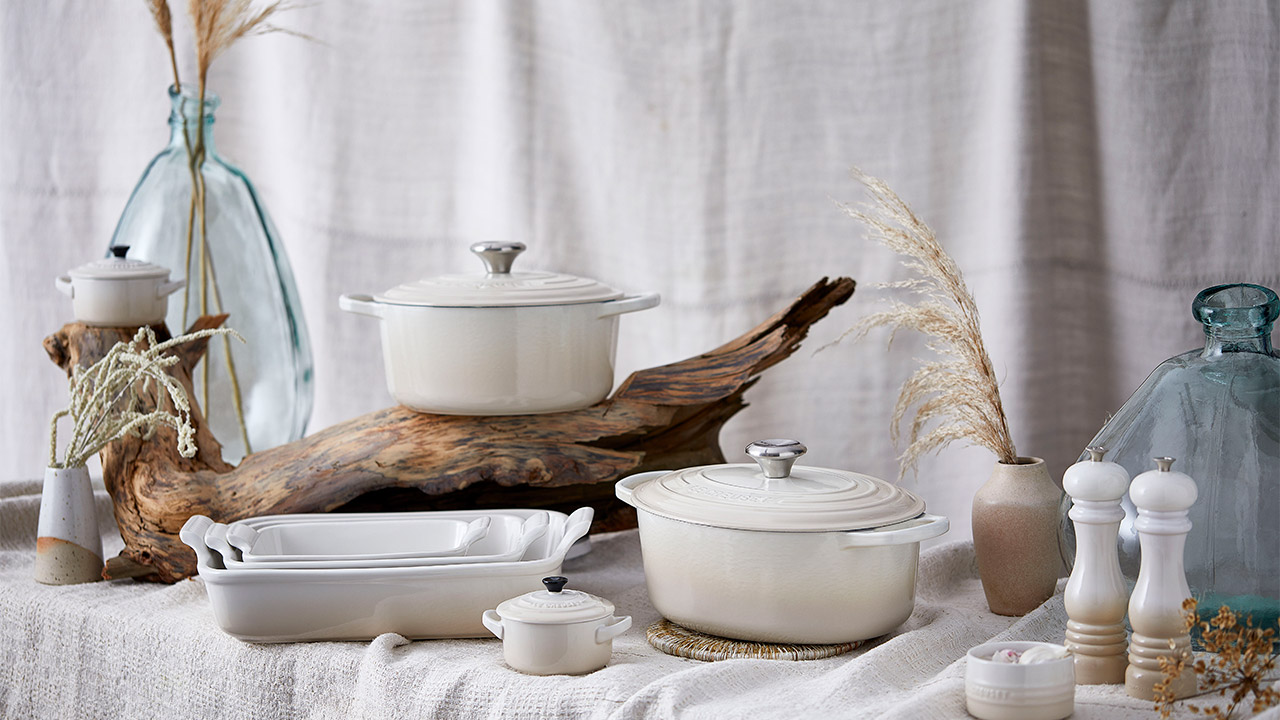 White ceramic cookware and decorative vases on a textured cloth backdrop with driftwood accents.

