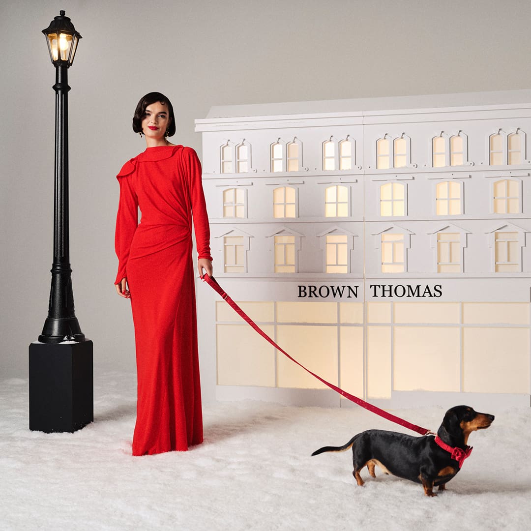 A woman in a red dress stands with a dachshund on a leash near a building facade labeled 'Brown Thomas.'