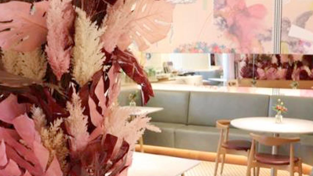 A cozy cafe interior featuring pink and white dried floral arrangements with round tables and green cushioned seating.