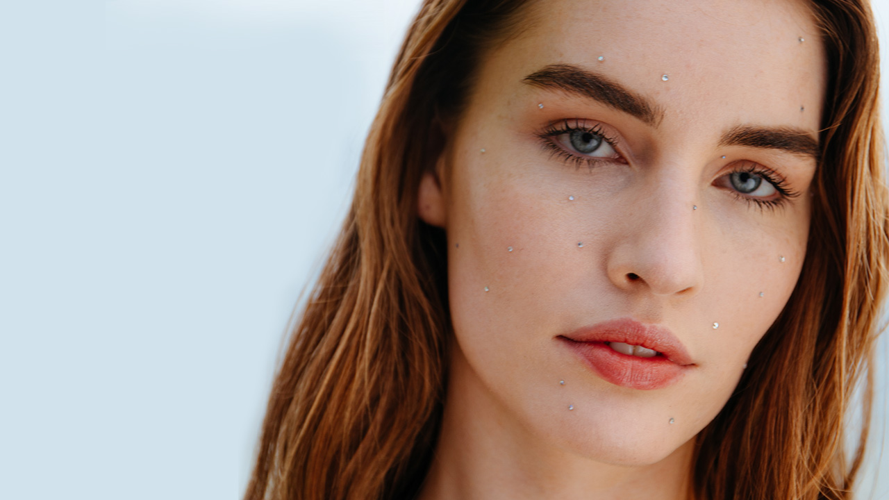A person with long hair and subtle makeup, wearing small facial embellishments, against a light background.