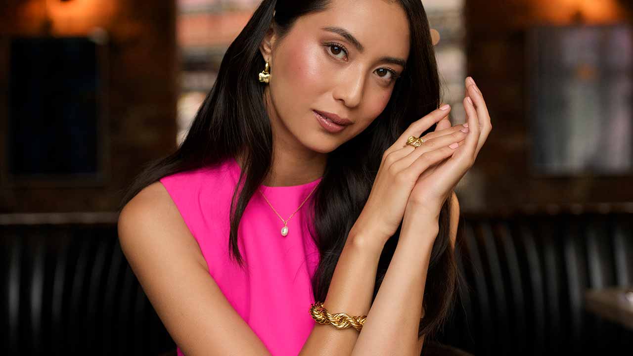 A woman in a pink top, adorned with gold jewelry, poses gracefully indoors.