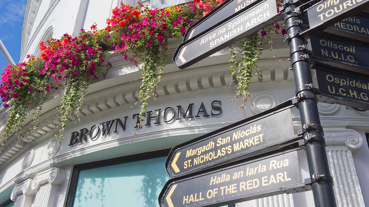 Street signs in front of a building with a 'Brown Thomas' sign and vibrant flowers hanging above.