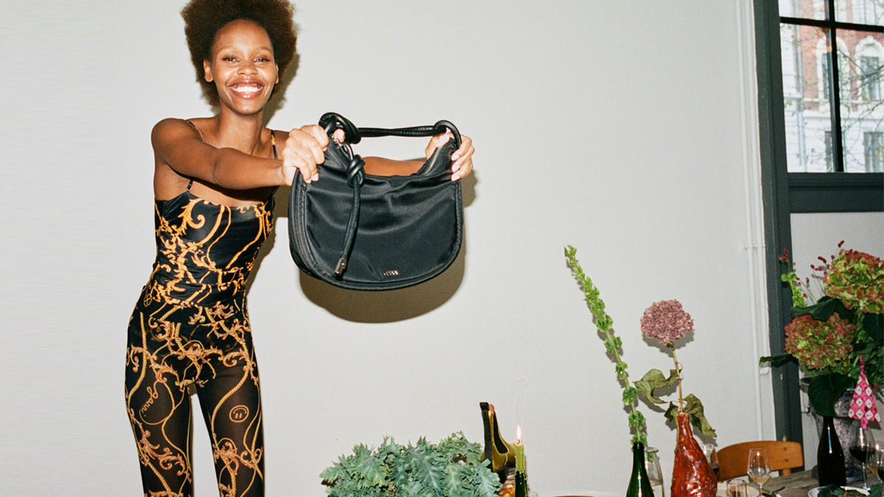 A smiling person in a patterned outfit holds up a black handbag in a room decorated with plants and flowers.