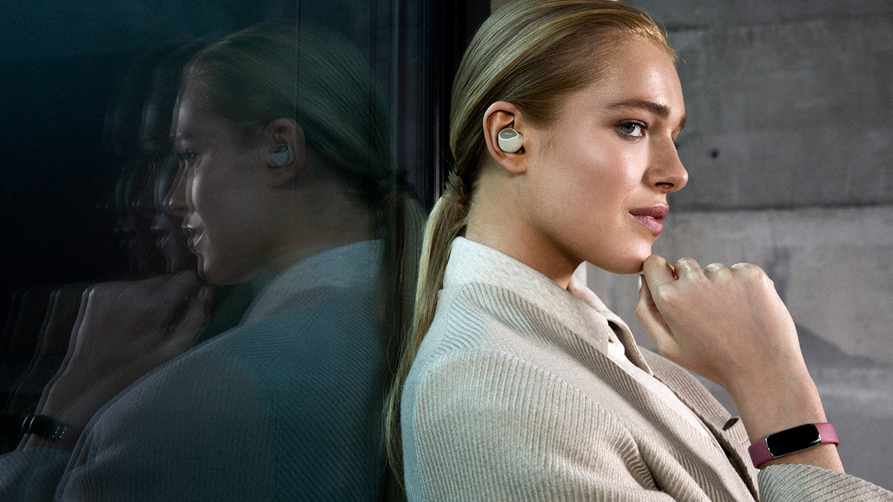 A person wearing wireless earbuds and a smart wrist device leans against a reflective surface.